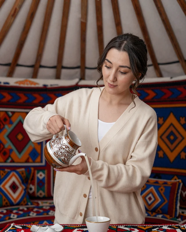 Kyrgyz woman inside a traditional yurt decorated with colorful felt carpets, serving traditional beverages